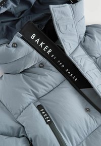 Veste matelassée gris clair avec une texture lisse et brillante, dotée d'une étiquette noire avec "BAKER by TED BAKER" et de poches zippées.