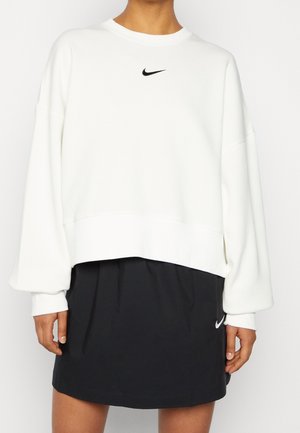 Osoba ubrana w białą, oversize'ową bluzę i czarną spódnicę, obie z małymi logo Nike, stojąca na tle jednolitego, białego tła.