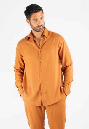 Chemise à boutons à manches longues de couleur rouille, fabriquée dans un tissu texturé, offrant une coupe décontractée et un ourlet arrondi. Assortie à un pantalon correspondant.
