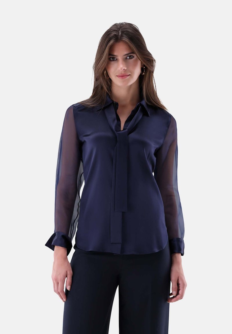 Navy Satinbluse mit durchsichtigen langen Ärmeln, Kragen und passendem Binder. Mit abgerundetem Saum und schwarzen Manschetten. Elegante, glatte Textur.