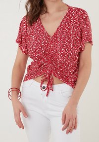 Blouse florale rouge à manches courtes, décolleté en V, avec un détail de nœud froncé à l'avant, associée à un jean blanc et des bracelets rouges.