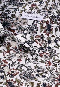 Chemise blanche avec motif floral multicolore en rouge, noir et vert, dotée de boutons blancs et d'une étiquette "seidensticker" visible sur le col.