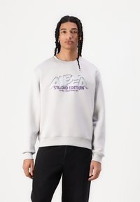 Sweat-shirt gris clair avec des poignets et un ourlet c�ôtelés, arborant un logo réfléchissant "ALPHA STUDIO EDITION" en violet et argent à l'avant.