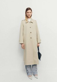 Trench coat bej deschis, cu guler, închidere cu nasturi și buzunare laterale; purtat peste blugi lejeri de culoare bleu deschis și pantofi albi cu vârf ascuțit.