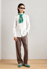 Witte overhemd met knopen, groene gestippelde sjaal, bruine broek en groene sneakers met witte strepen. Accessoires zijn een zonnebril en een ring.