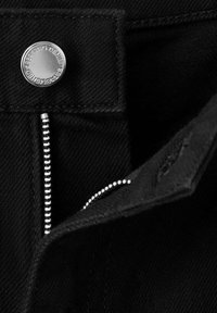 Gros plan d'un tissu en jean noir avec une fermeture éclair argentée non fermée et un bouton argenté portant l'inscription "1989 KING DENIM ESTABLISHED."