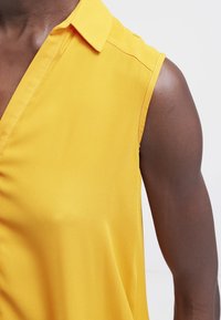 Žlutá blůza s krátkým rukávem a límečkem, vyrobená z hladkého materiálu. Má design s drapovaným předním dílem.