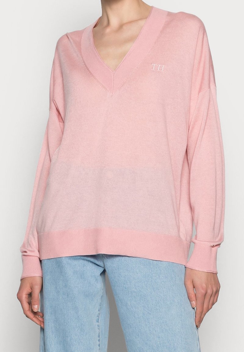 Femme portant un pull ample rose à encolure en V avec des poignets et un ourlet côtelés, associé à un jean denim bleu clair.