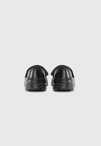 Zapatos negros con un acabado de cuero suave, parte trasera redondeada, correa elástica en el talón y suela de goma texturizada.
