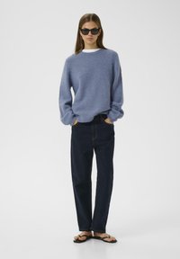 Helles blaues, flauschiges Sweatshirt mit langen Ärmeln, kombiniert mit dunkelblauen, geraden Jeans und schwarzen Flip-Flops. Weißes Unterhemd ist am Halsansatz sichtbar.