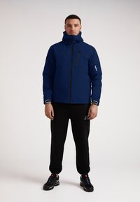 Mørkeblå isoleret jakke med høj krave og lynlåslommer, i kombination med sorte sweatpants. Har ensfarvede elementer og en afslappet pasform.