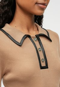 Καφέ ραφωτό τοπ με μαύρο δερμάτινο γιακά και πασπαρτού με τρία κουμπιά σε χρυσό χρώμα. Κοντινό πλάνο του λαιμού και της υφής του υφάσματος.