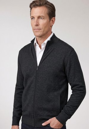 Mann trägt eine dunkelgraue Jacke mit Reißverschluss über einem weißen Hemd, steht vor einem neutralen hellen Hintergrund, mit einer Hand in der Tasche.