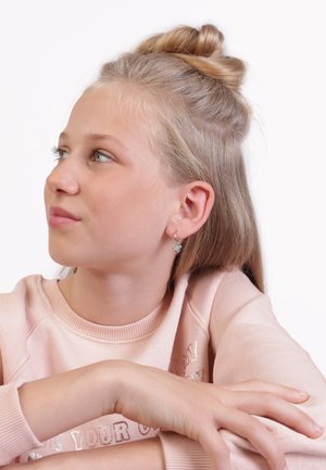 Zilveren hartvormige oorbellen hangen van doorboorde oren. Het onderwerp draagt een lichtroze sweatshirt met een ronde hals en geribbelde manchetten.
