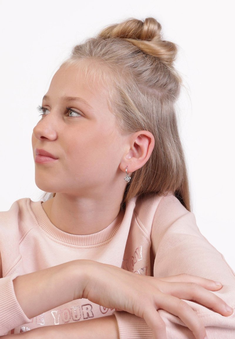Silberne herzförmige Ohrringe hängen von durchstochenen Ohren. Das Modell trägt einen hellrosa Sweatshirt mit Rundhalsausschnitt und gerippte Bündchen.