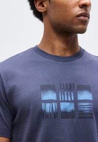 Man draagt een blauw T-shirt met een afbeelding van drie abstracte blauwe en zwarte rechthoekige patronen op de borst.