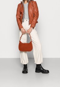 Schultertasche aus Rostleder mit abgerundeter Form und tonalem Zierstich. Kombiniert mit einem grauen Oberteil, brauner Lederjacke, cremefarbenen Hosen und schwarzen Stiefeln.