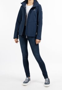 Navyblå softshelljacka med hög krage, framzip och två fickor med dragkedja på sidorna. Har ankarlogotyp på bröstet, tillsammans med blå jeans och sneakers.