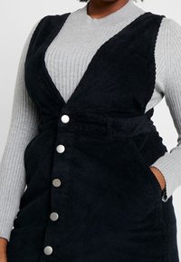 Schwarzes Cordrobenkleid mit V-Ausschnitt, silbernen Knopfdetails und seitlichen Taschen, übereinander getragen mit einem hellgrauen, gerippten langärmeligen Oberteil.