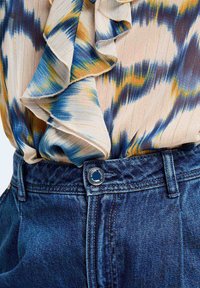 Trykt bluse med blå, gul og beige mønstre med volanger. Kombinert med mørkeblå denimjeans med knappelukking.