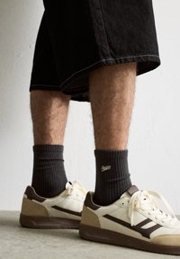 Sneakers med en cremet overdel, brune accenter og en tekstureret sål. Sættes sammen med mørke ribbede sokker og sorte shorts.