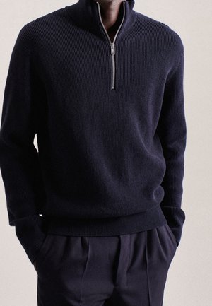 Mężczyzna w granatowym prążkowanym swetrze z półzamkiem przy kołnierzu i ciemnych spodniach, ręce w kieszeniach, na neutralnym tle.