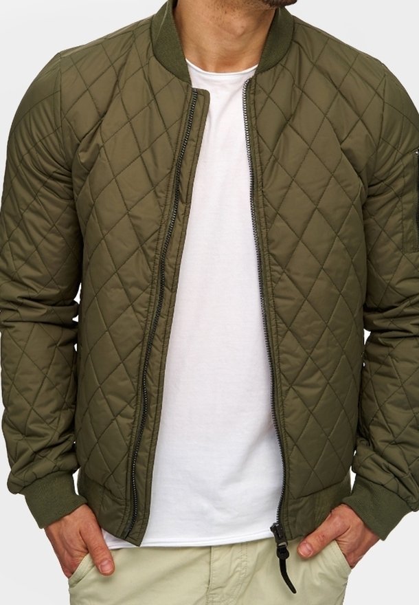 Olivgrüne gesteppte Bomberjacke mit gerippten Bündchen und Kragen, ausgestattet mit einem Frontreißverschluss und Seitentaschen, über einem weißen Hemd getragen.