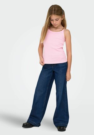 Ragazza giovane con lunghi capelli biondi, che indossa una canottiera rosa, jeans blu a gamba larga, scarpe nere e una molletta per capelli, in piedi e guarda verso il basso.