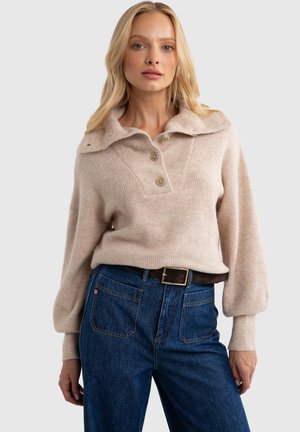 Beżowy sweter dziany z szerokim kołnierzem, trzema guzikami z przodu i długimi, balonowymi rękawami. Noszony na ciemnoniebieskich, wysoko osadzonych dżinsach.