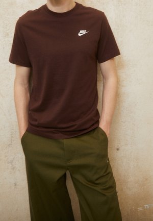 Persona vistiendo una camiseta Nike de color marrón oscuro y pantalones verde oliva, de pie contra una pared clara con textura, con las manos en los bolsillos.