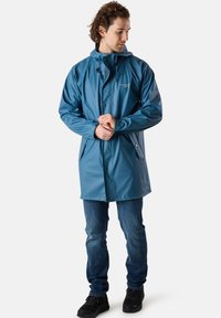 Blå vattentät jacka med huva, tryckknappar och sidofickor, tillverkad av slät, glänsande material. Buren med jeans och svarta skor.