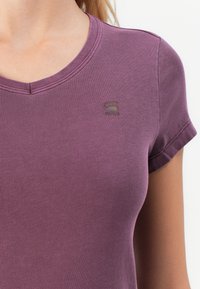 Nahaufnahme einer Person, die ein mauvefarbenes T-Shirt mit kurzen Ärmeln trägt. Auf der Brust, in der Nähe des Schlüsselbeins, befindet sich ein kleines, dunkles Logo.