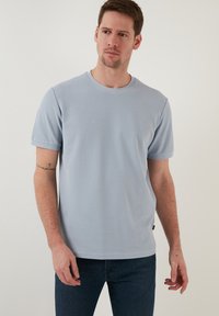 Tricou scurt cu mânecă din material texturat, de culoare albastru deschis, cu guler rotund și croială ajustată. Purtat cu blugi negri.