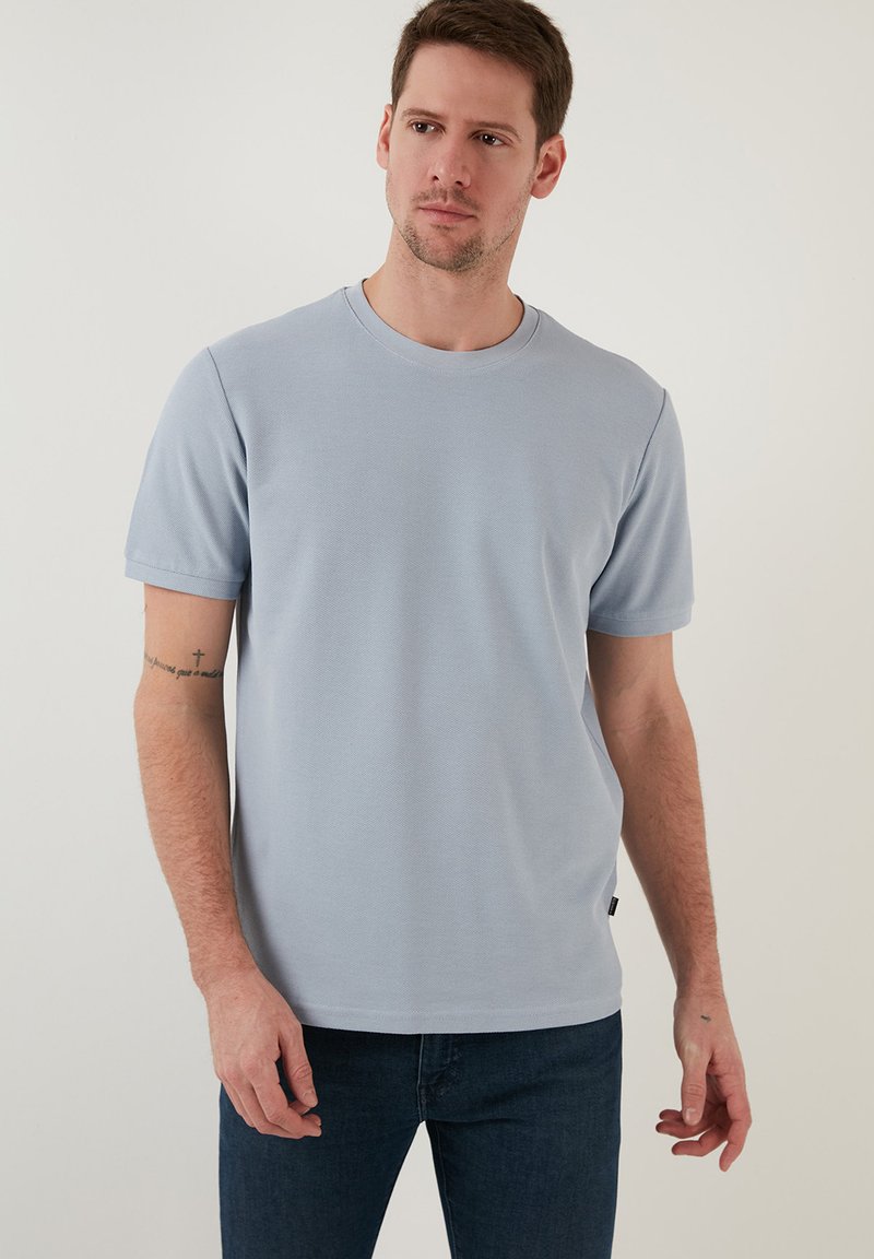 Tricou scurt cu mânecă din material texturat, de culoare albastru deschis, cu guler rotund și croială ajustată. Purtat cu blugi negri.