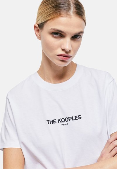 Baltās kokvilnas T-veida blūze ar melnu "THE KOOPLES PARIS" logo, kas centrēta krūtīs. Īsas piedurknes un apaļa kakla izgriezums.