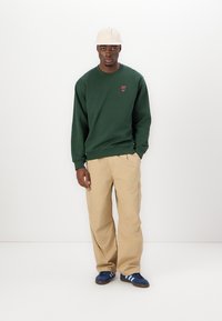 Green sweatshirt with a small red logo, beige cargo pants, and dark blue sneakers with white stripes. Model wears a light beige cap.