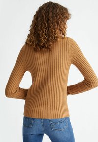 Kobieta z kręconymi włosami nosząca sweter w kolorze khaki i niebieskie dżinsy, stojąca z rękami na biodrach, odwrócona plecami.