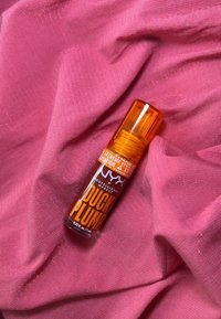 NYX Duck Plump lipproduct in een doorzichtige oranje fles met een spraymondje, geplaatst op een textuur van roze stof.