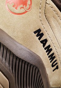 Beżowe zamszowe buty z czarnym logo "MAMMUT" oraz teksturowaną brązową gumową podeszwą, charakteryzującą się wyraźnym wzorem rowków dla lepszej przyczepności.