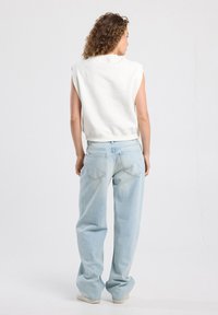 Mujer con cabello rizado que lleva una blusa sin mangas blanca y unos jeans sueltos de color azul claro, de pie de espaldas contra un fondo liso.