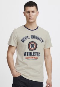 Bežinė marškinė su apvaliomis i�škirptėmis, tamsiai mėlynas apykaklė. Priekinė grafika turi ryškų tekstą "Dept. Varsity Athletic," su skydu ir oranžiniu akcentu.