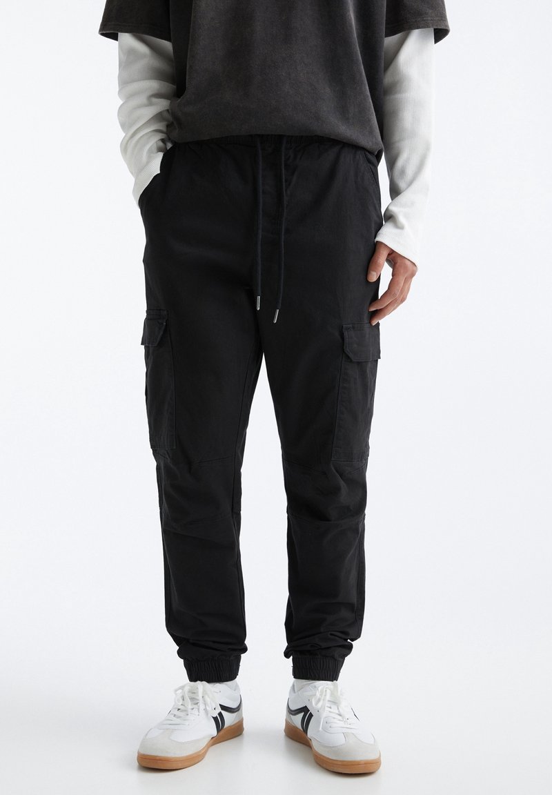 Schwarze Cargo-Hosen mit elastischen Bündchen, ausgestattet mit frontalen Taschen und einem Kordelzugbund. Getragen mit einem dunklen T-Shirt und einem weißen Langarmhemd.