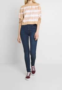 Ljusbrun kort sweatshirt med vitt tie-dye-mönster, kombinerad med mörkblå skinny jeans och röda sneakers. Slät textur, avslappnad design.