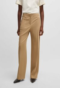 Beige tailored trousers with a smooth texture, wide legs, and side zip pockets. Paired with a light-colored top and pointed black shoes.