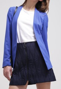 Blazer azul a medida con mangas largas sobre una camisa blanca, combinado con una falda negra ajustada que presenta un patrón de olas texturizado y cremallera.