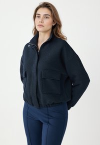 Vrouw in een marineblauwe gestructureerde vest met rits en grote voorzakken, gecombineerd met bijpassende marineblauwe broek, staand tegen een witte achtergrond.