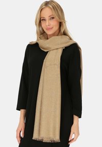 Beżowy szalik z kwiecistym wzorem i subtelnym połyskiem, z frędzlami na końcach. Nosi się luźno na czarnym topie. Miękka tekstura.