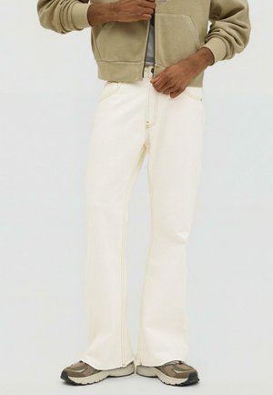 Persona che indossa una felpa con cappuccio beige con cerniera, jeans dritti color crema e sneakers beige e grigie, che si sistema la vita dei jeans.