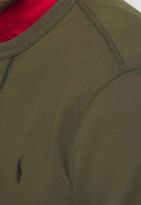 Olivgrön sweatshirt med röd insida, med rund halsringning, insydda ärmar och en liten svart logotyp på bröstet. Mjuk textur.