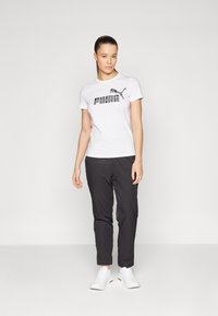 White cotton t-shirt with black 'PUMA' logo, paired with dark trousers. Model wears white athletic shoes; simple background, neutral tones.